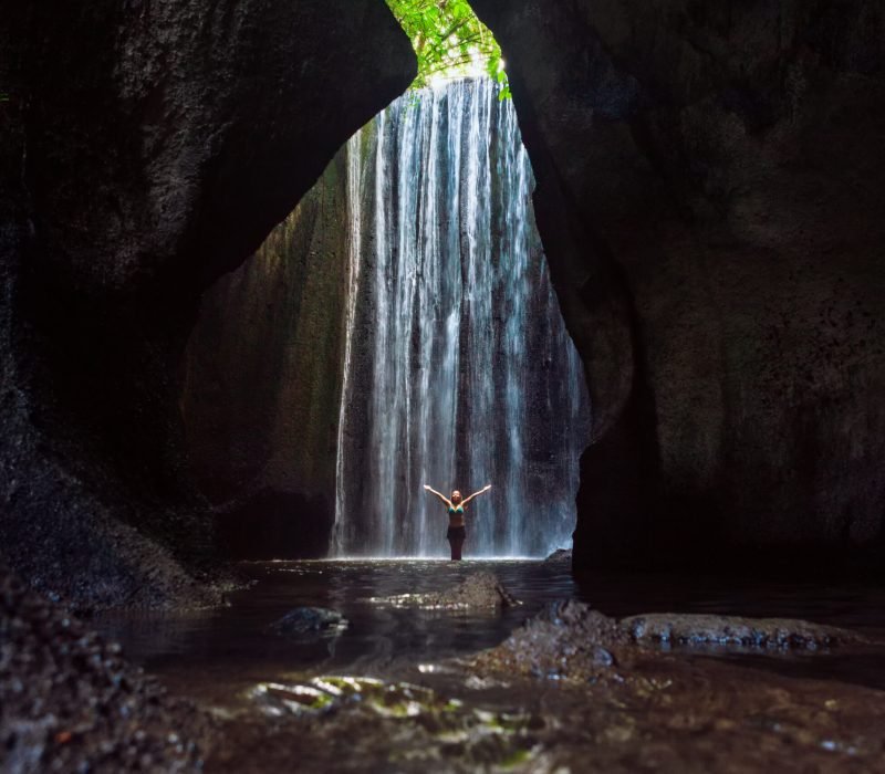 Woman stand in underground cave pool under falling fresh water of Tukad Cepung waterfall. Nature day tour, hiking activity adventure and fun at family tourist camp on summer vacation in Bali island