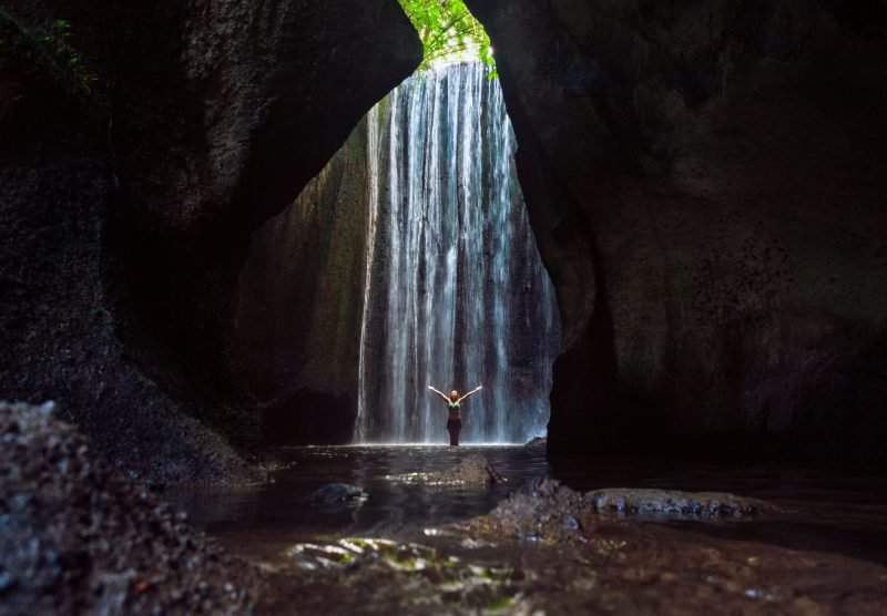 Woman stand in underground cave pool under falling fresh water of Tukad Cepung waterfall. Nature day tour, hiking activity adventure and fun at family tourist camp on summer vacation in Bali island